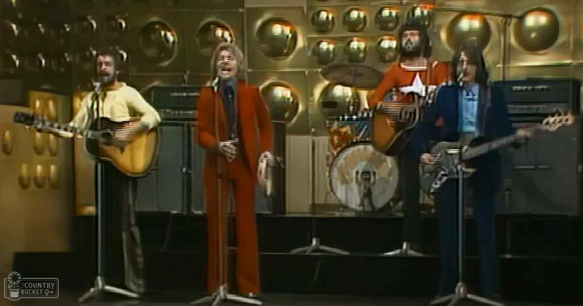 Adult male singer in a long-sleeved shirt, tie, and dark pants holding an acoustic guitar; adult male in a bright red suit and white shirt, adult female in a dark blue dress or jumpsuit, all performing on stage with futuristic reflective panels in the background.