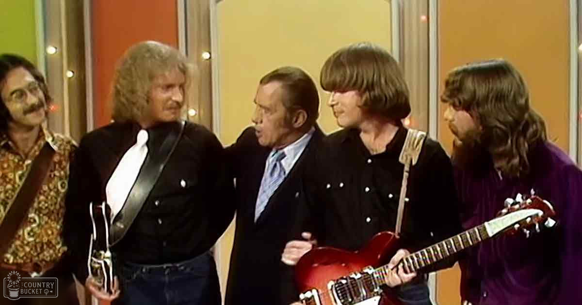A musical group performing in a television studio or stage setting with vertical panel backdrops under artificial lighting.