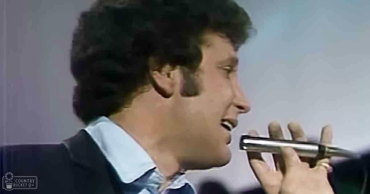 A young adult male singer in a dark suit jacket with a white shirt, holding a metallic microphone on stage at Madly Odd.