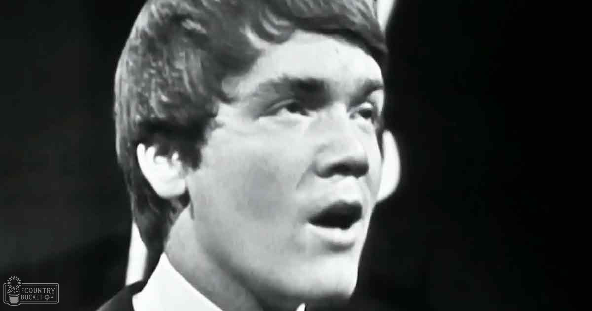 A young adult male in a formal or semi-formal white collared shirt with dark jacket, standing indoors under dramatic lighting, possibly on stage.