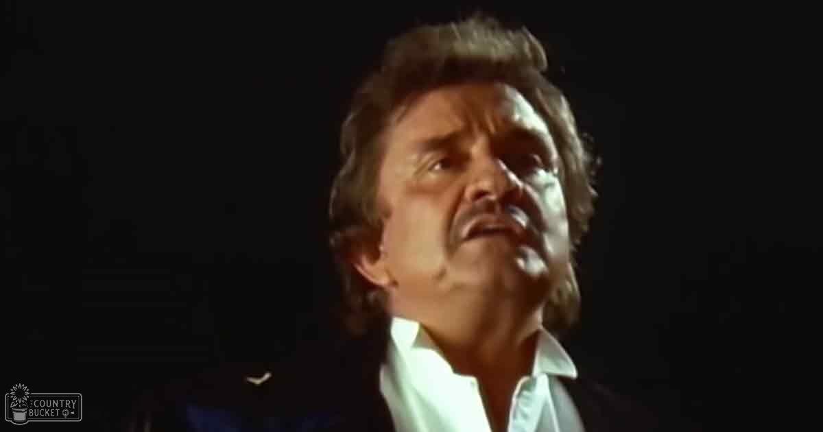 A middle-aged adult male in a relaxed, casual, collared shirt with long sleeves, standing indoors under dark lighting conditions typical of performances or recordings.