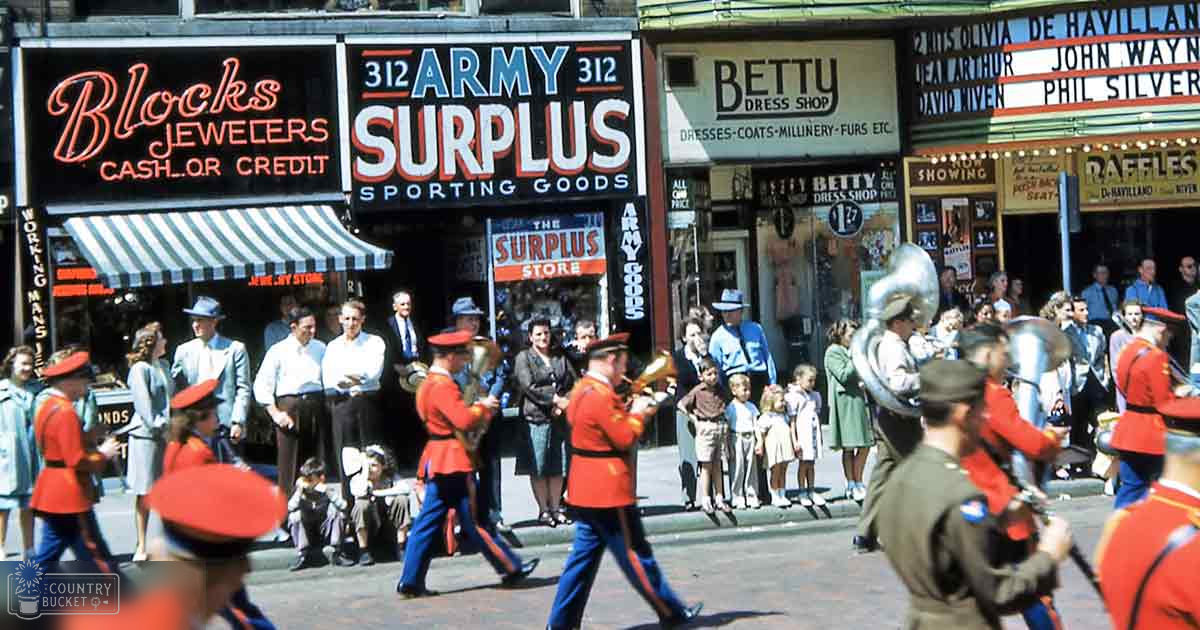 A parade in an urban setting with marching bands and a diverse crowd of adults and children dressed in mid-20th century fashion.
