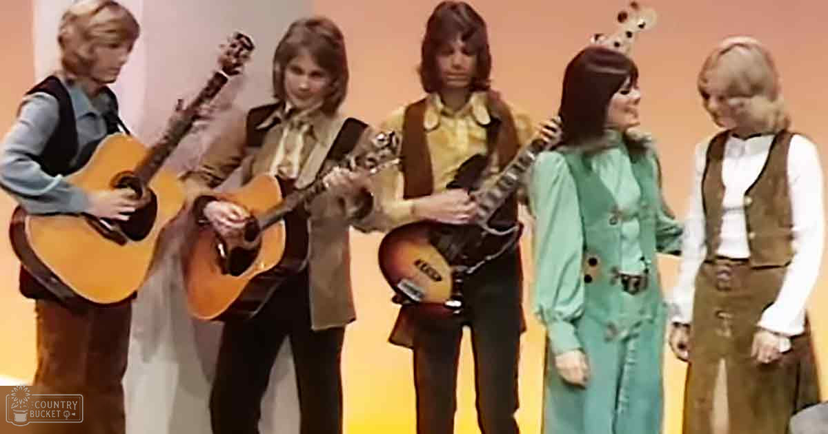 Young adult musicians in a 1970s-style musical performance group indoors with plain backdrop.