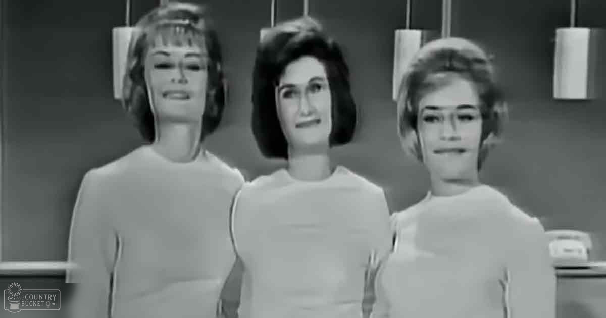 Three young adult females in a kitchen setting wearing 1960s-style bob-cut garments with smooth cotton or polyester fabric, hanging cylindrical light fixtures in the background.