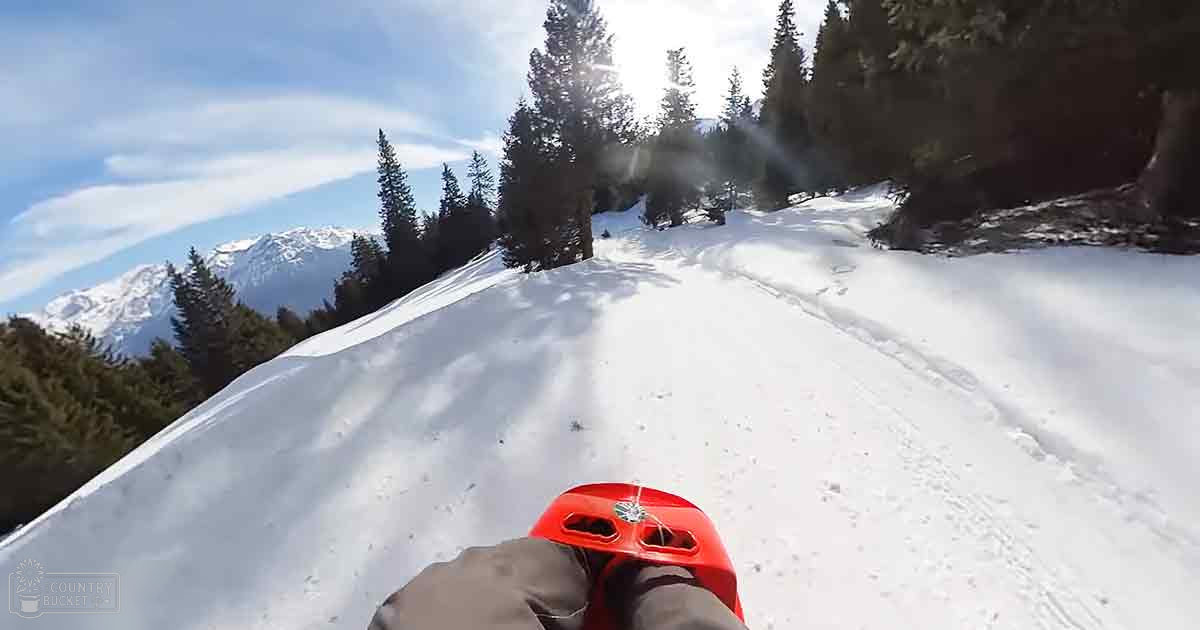 A young adult or middle-aged male sledding down a snowy slope in a mountainous region with coniferous trees.