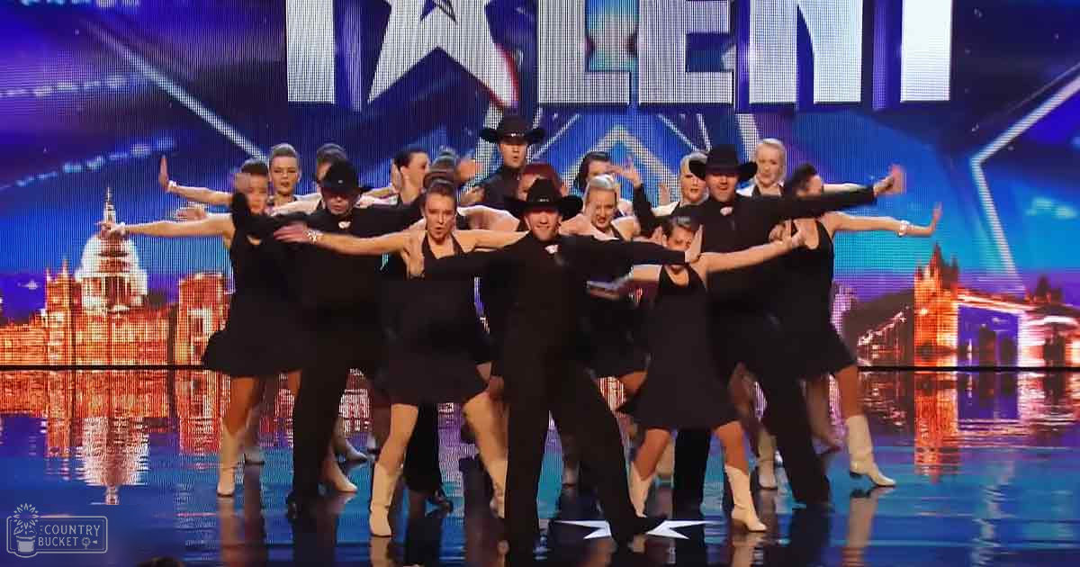 A dance group performing at a talent show with young adult females in black dresses and middle-aged men in black suits, cowboy hats, and white boots under vibrant stage lighting.