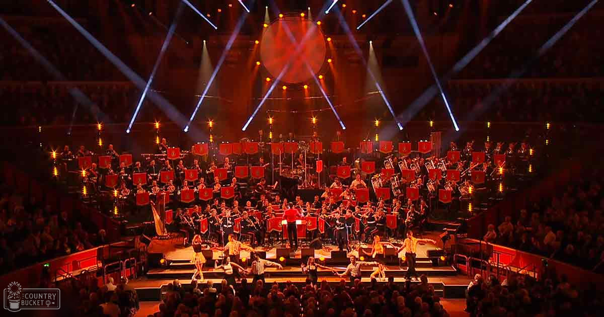 A large orchestra performing in an indoor arena or concert hall with tiered seating filled with spectators, stage lights illuminating red podiums and musicians in dark uniforms with red accents and white collars. The setting suggests a formal evening performance.