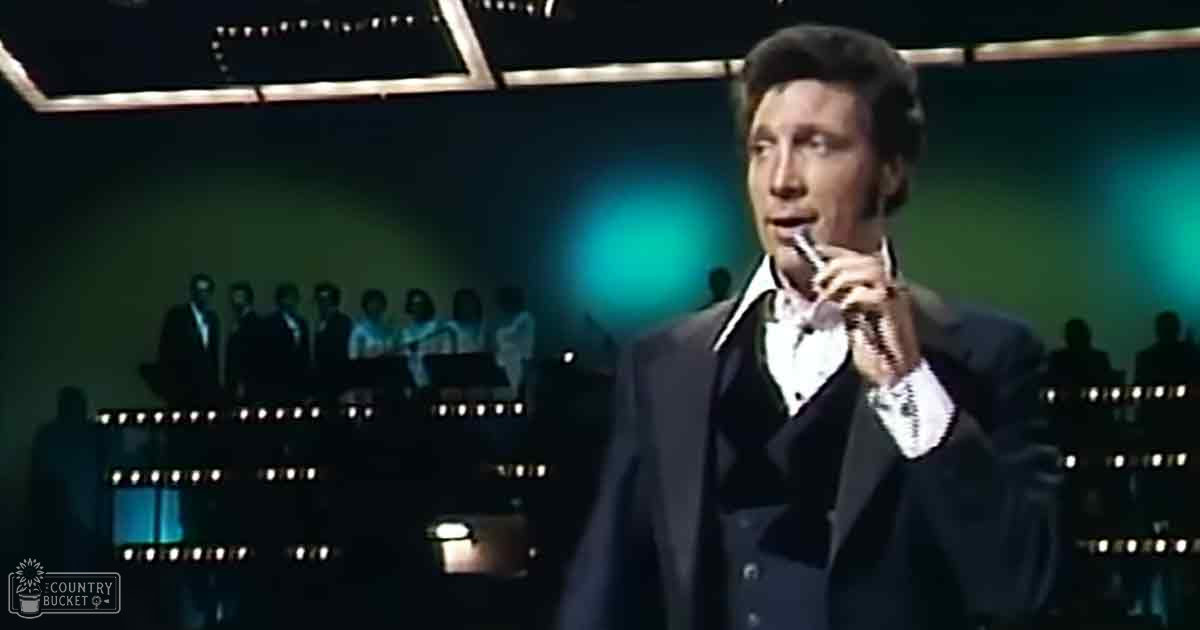 An adult male singer in a dark mid-20th-century style suit with white shirt and black bow tie, performing on stage against silhouetted figures in formal attire under evening lighting.