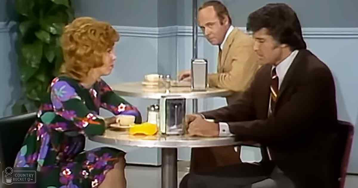 A woman in a floral dress converses with two men in formal attire at an indoor setting, possibly during a talk show or interview.