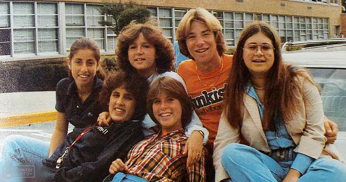 A group of children and an adult female standing in front of a multi-story building, possibly a school or community center. The setting appears to be outdoors with no snow on the ground.