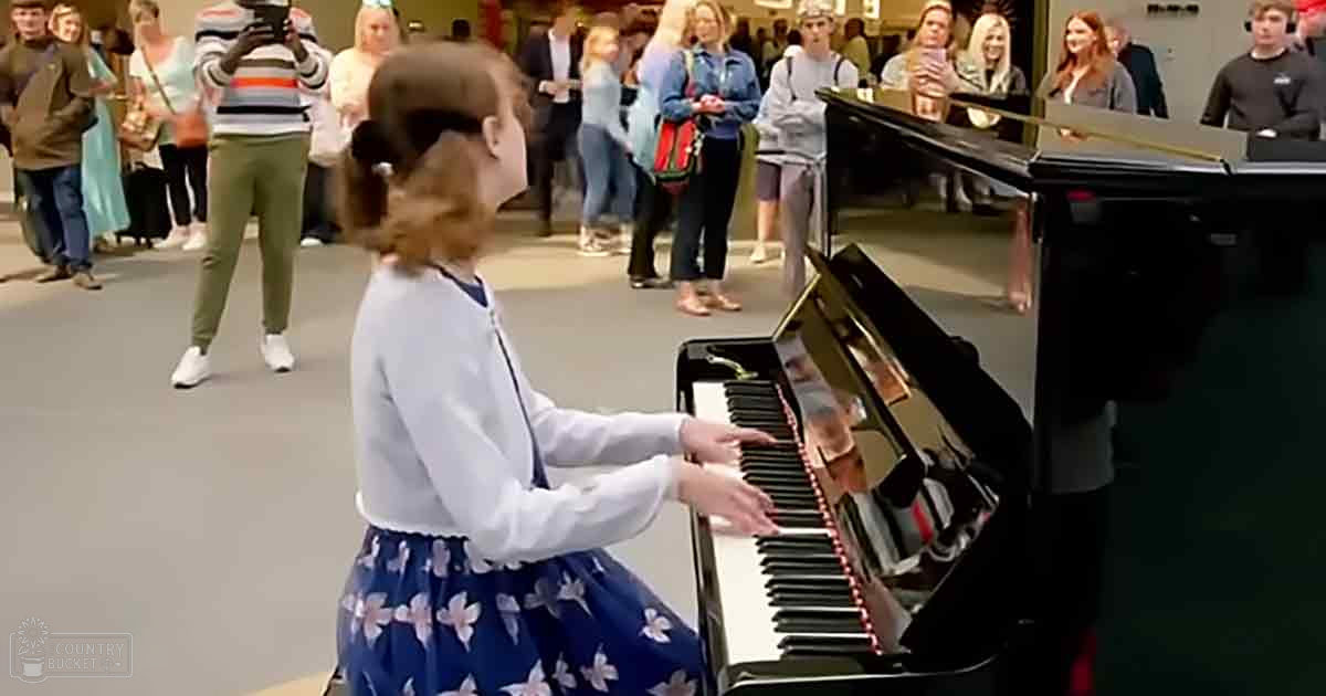 A young child wearing a white long-sleeve top with dark trim and blue floral skirt plays piano amidst onlookers in an indoor public space.