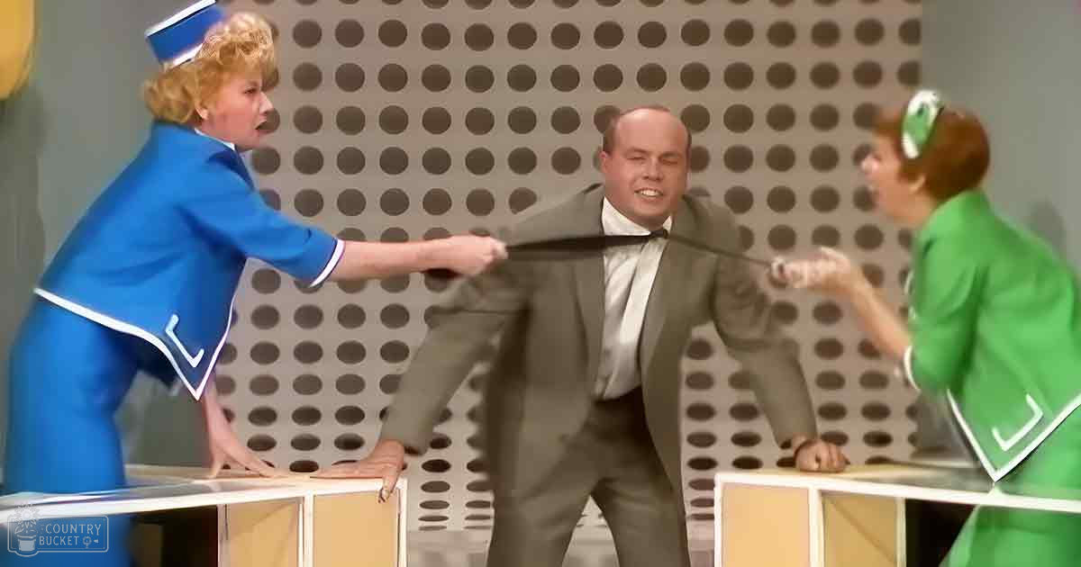 A comedic scene from a television show or sketch comedy program, possibly set in a studio environment with three individuals dressed as flight attendants and a man in formal attire standing behind white cubicles resembling game show segments.