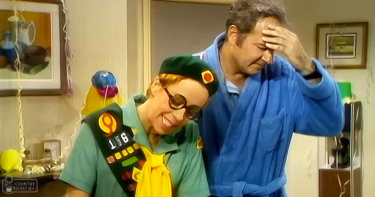 Indoor home scene with two individuals; child/teen male wears green shirt and yellow tie with badges, middle-aged male in blue bathrobe; shelves with kitchenware, framed pictures on wall; setting suggests kitchen or dining area.