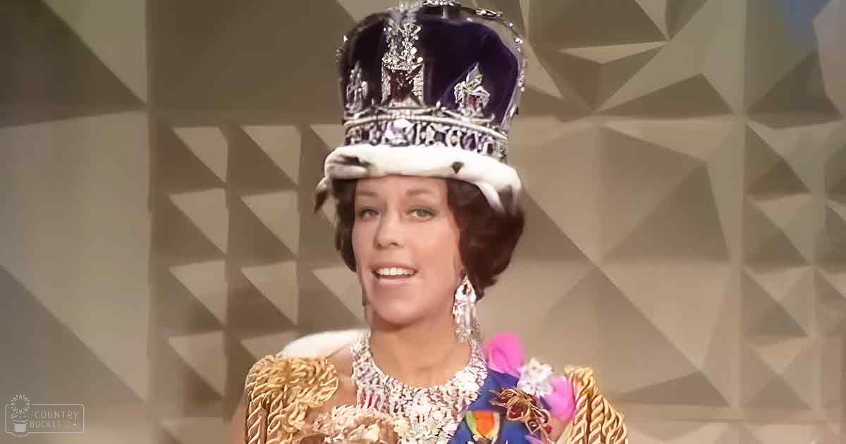 A woman in a luxurious, elaborate gown with intricate gold embroidery and vibrant colors like purple, pink, and green, wearing an ornate crown and dangling earrings, seated indoors with geometric patterns.