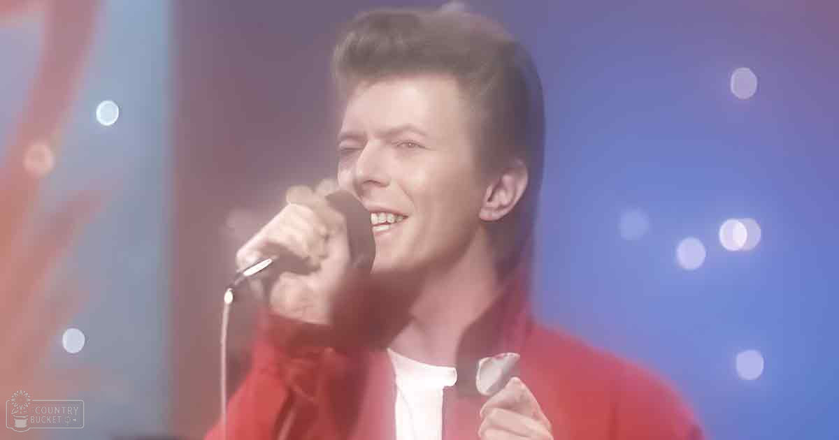 A young adult male singer in a relaxed fit red jacket over a white shirt with 'Madly Odd' text on stage, illuminated by blue stage lights.