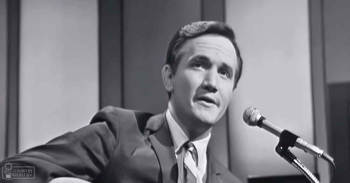 A middle-aged adult male in a tailored dark suit with a collared shirt and tie, standing indoors near a microphone setup, possibly in a recording studio or radio broadcast environment.