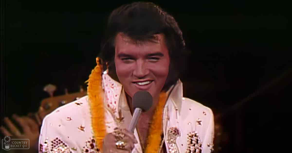 An adult male singer in a white sequined jacket with gold accents, performing on stage under dramatic lighting. He holds a microphone, wearing a form-fitting and embellished garment.