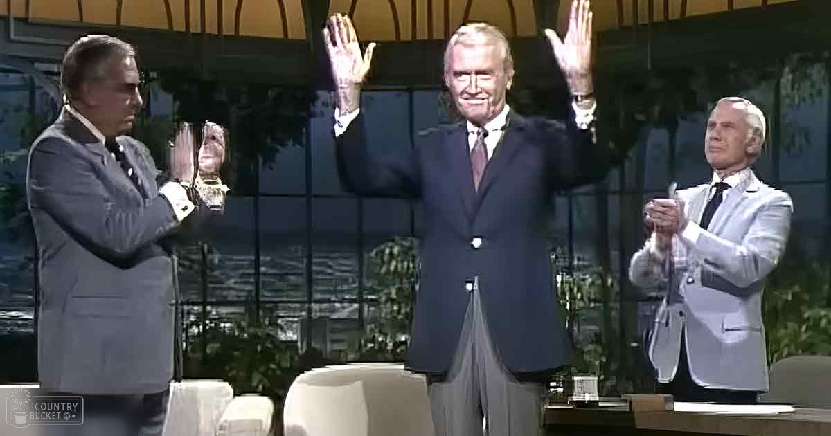 Three adult males, dressed formally for a talk show event, seated indoors with natural daylight illuminating a backdrop of lush greenery.