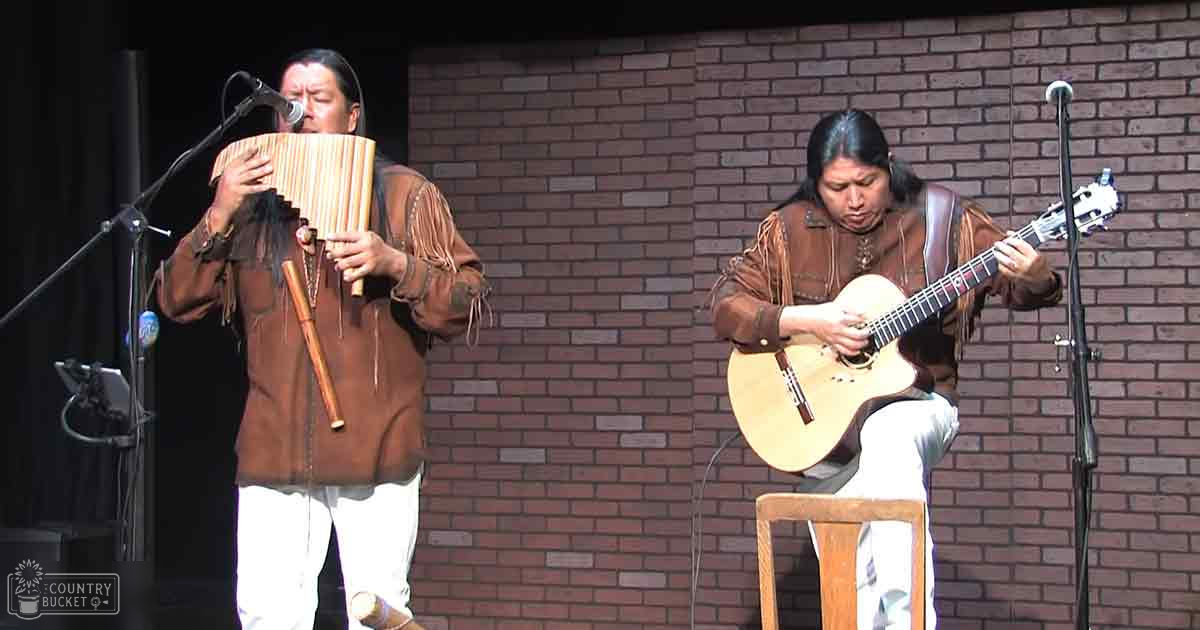 Musical performance with two adults, one male singer wearing a brown leather jacket with fringe details and white pants; the other female musician in similar attire but darker tones, seated with a guitar strap. Setting appears to be an indoor venue with brick walls, wooden furniture, and bright lighting.