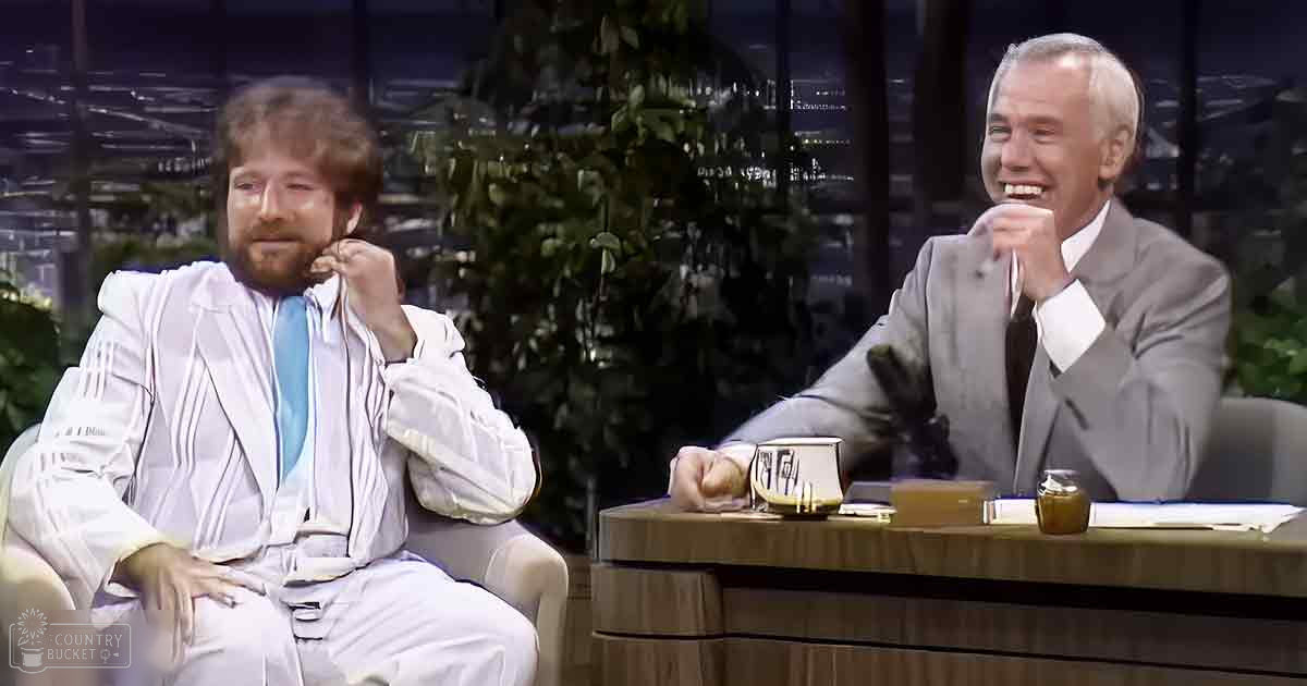 Talk show host and guest in an indoor studio setting with wooden furniture, potted plants, and a mug on a desk at night.