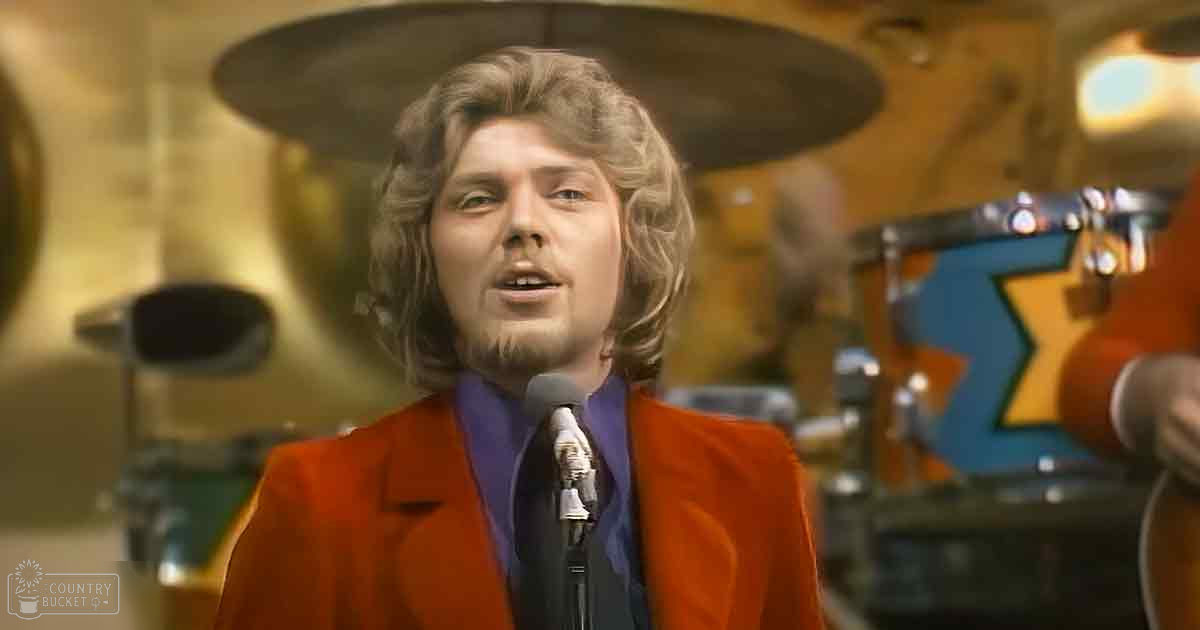 An adult male performer in a music venue, wearing an orange blazer and purple shirt with 'Madly Odd' text, surrounded by musical instruments under warm artificial lighting.