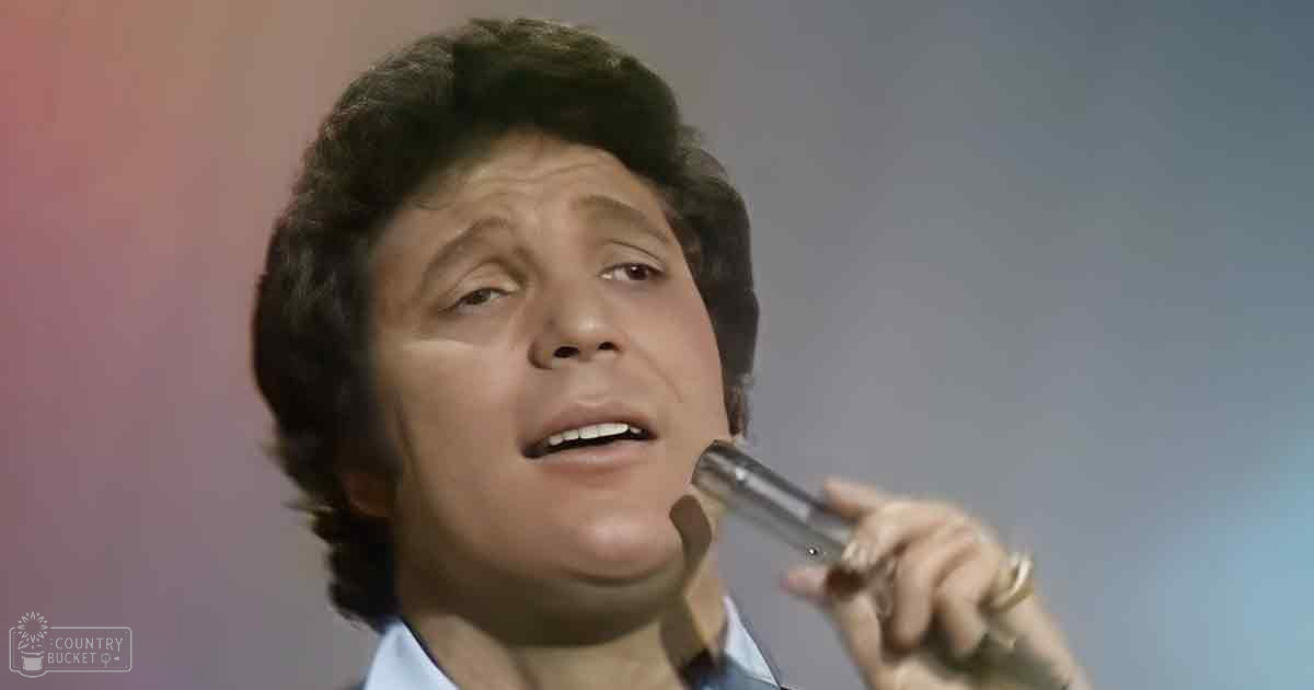 A middle-aged adult male singer wearing a light blue collared shirt with short sleeves, holding a microphone on an indoor stage or studio background.