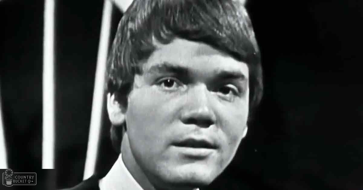 Young adult male wearing formal attire indoors, possibly at a formal event; dark wooden chair visible in the background.
