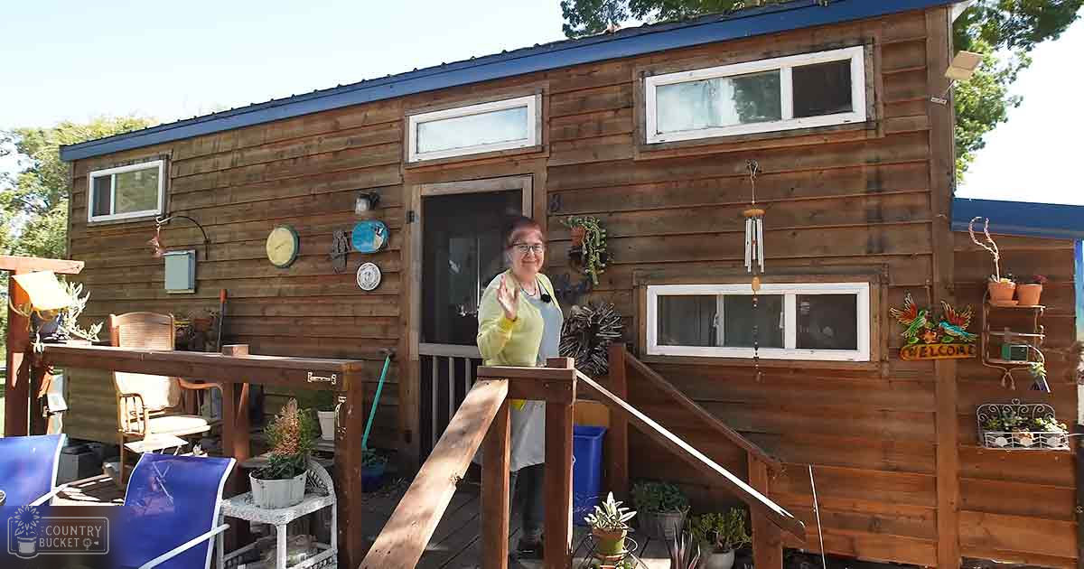 A middle-aged female wearing a light green long-sleeved top with white undershirt stands on a wooden deck in a residential area surrounded by outdoor furniture and greenery, likely spring or summer day.