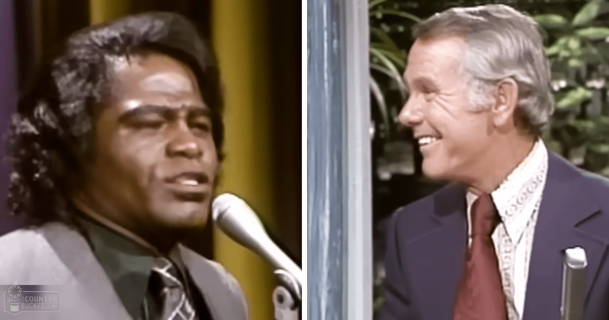 A male singer wearing a dark suit jacket and patterned shirt sings into a microphone while another middle-aged man, dressed in a white shirt with a patterned tie, appears as an interviewer or host against a backdrop of stage lighting and blurred greenery.