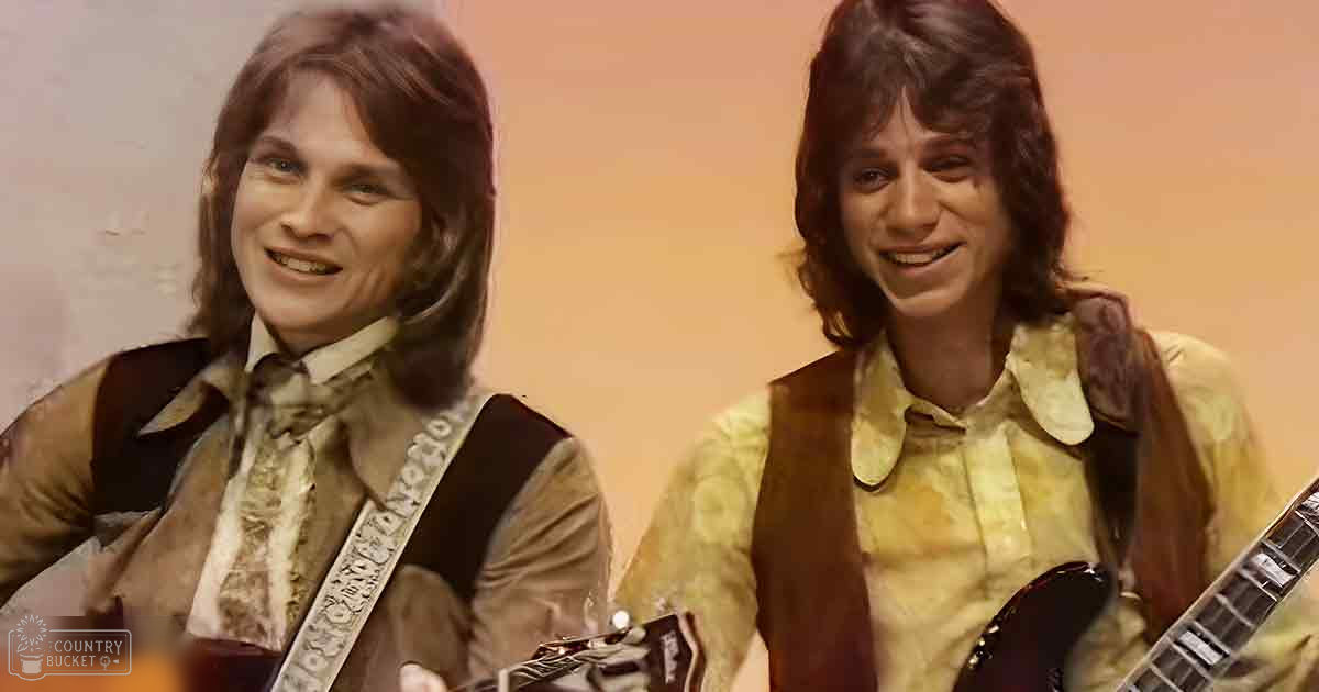 Two young adult males in a studio setting with guitars, wearing vintage-style clothing from the late 1960s to early 1970s.
