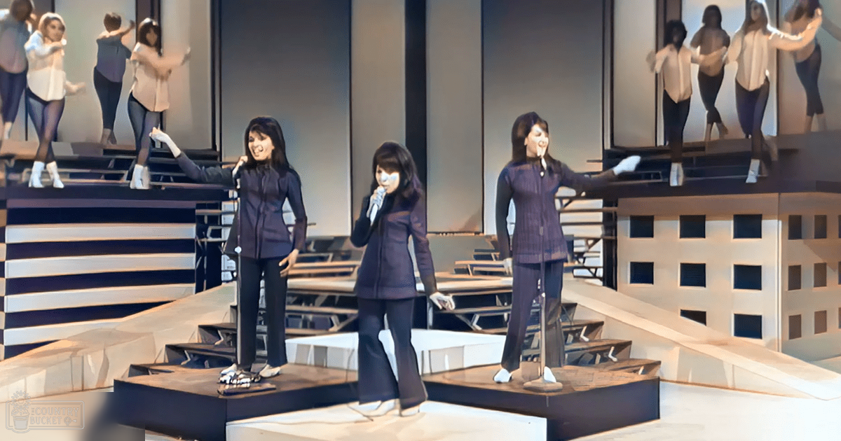 An indoor performance scene with three adult female performers on a stage-like area, displaying large screens behind them.