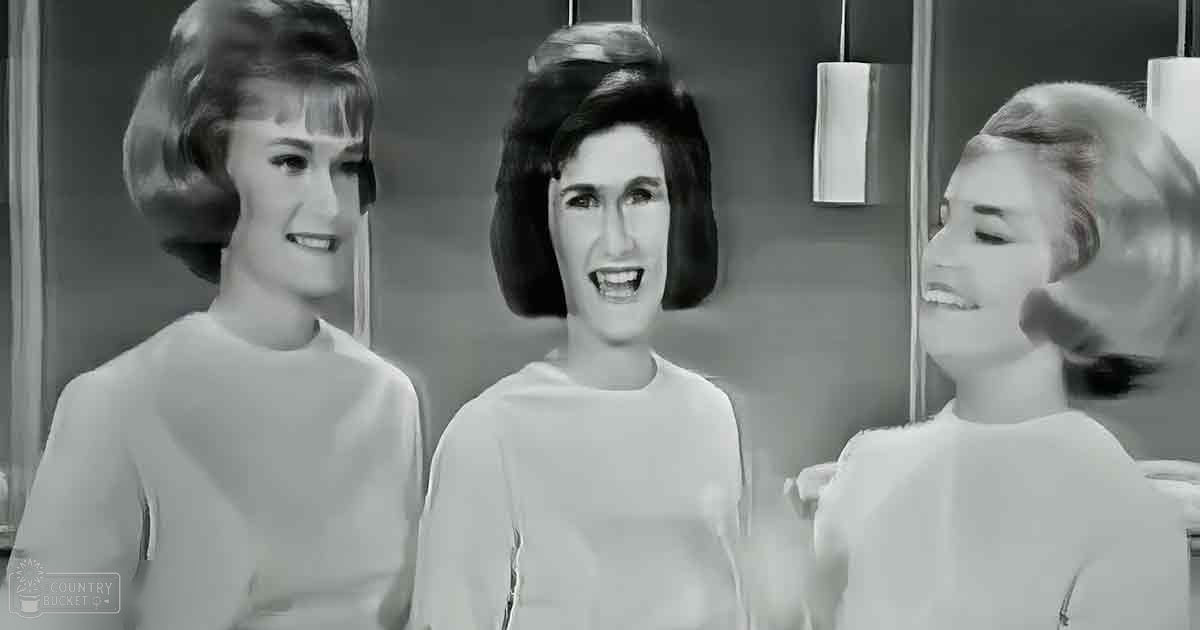 Three young adult women standing indoors, dressed in light-colored long-sleeved tops with simple cuts and necklines, possibly from a film or television show; setting appears to be an office or studio environment.