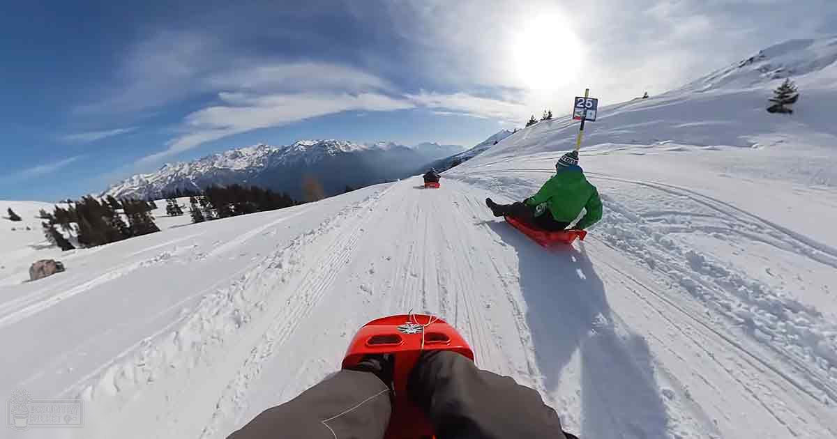 Young adult males sledding down a snowy slope in a mountainous area, wearing waterproof jackets during winter with bright daylight.