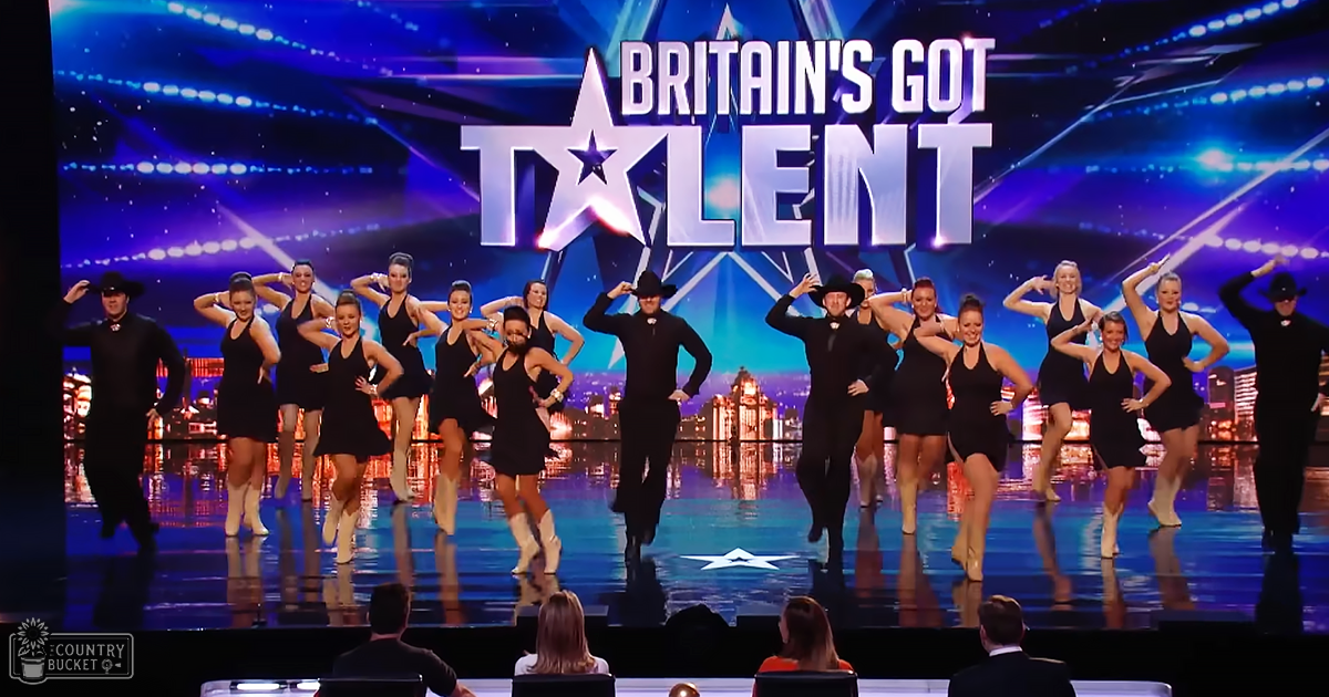 A dance group, mostly adult females wearing black sleeveless dresses with white knee-high boots, performing on a brightly lit stage at 'Britain's Got Talent' inside an indoor arena.
