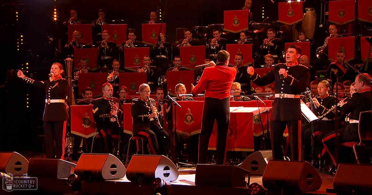 A military band performing at an indoor concert hall with red-accented uniforms and stage lighting equipment.