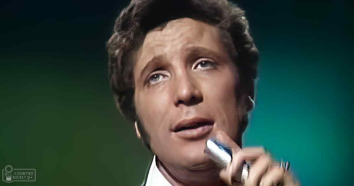 An adult male performer wearing a white shirt with a collar, holding a microphone on stage with a solid green backdrop.