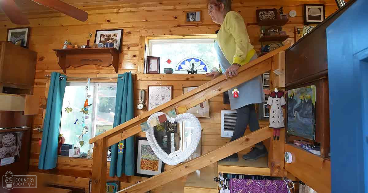 An older adult female wearing a light yellow cardigan over a blue top in a cabin with wooden walls, blue curtains, and various wall decorations.