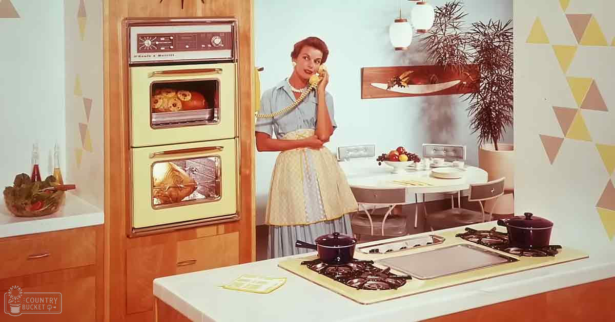 An adult female in a kitchen setting, wearing a light blue dress with a yellow apron, holding a telephone receiver.