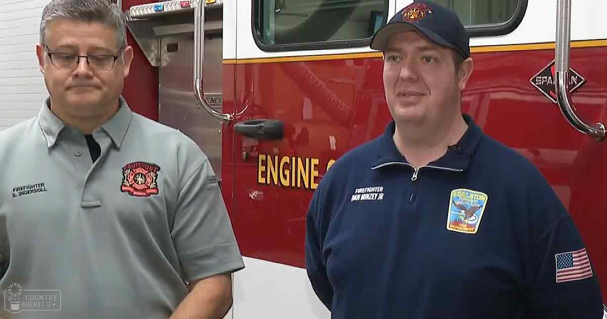 Two firefighters in front of a red fire truck with 'ENGINE' written on it, wearing gray and navy blue shirts with 'FIREFIGHTER' patches, standing outdoors at what appears to be a fire station during the daytime.