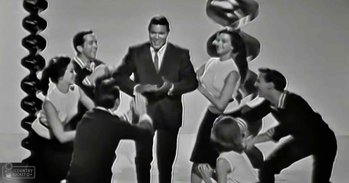 A group performance scene from the mid-20th century with a man in dark formal attire at center stage and other performers around him, set against a plain indoor backdrop.