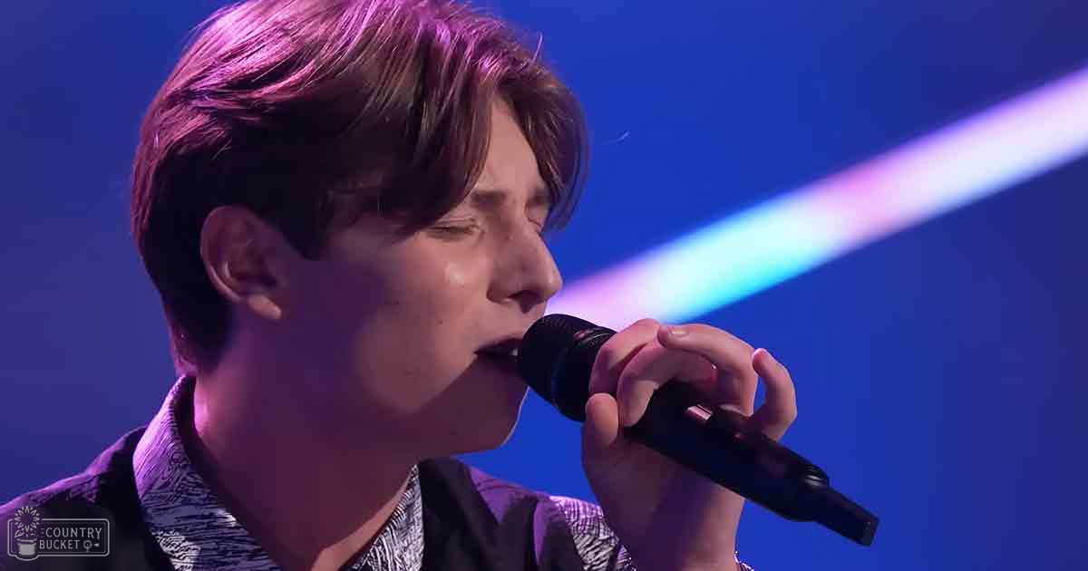 A young adult male singer wearing a button-up shirt with an abstract geometric print, singing into a microphone under stage lighting in a performance setting.
