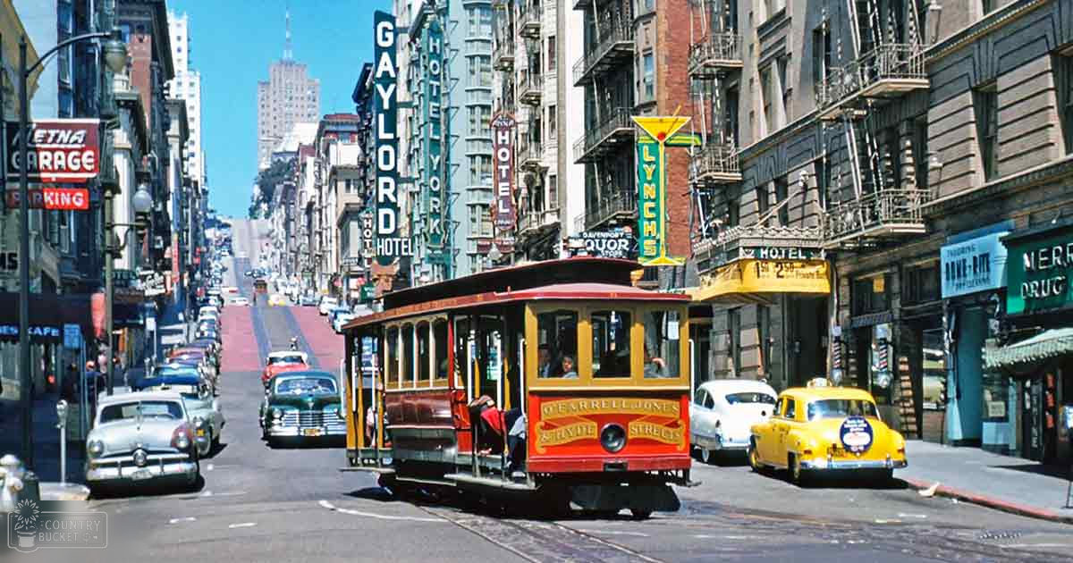 San Francisco's bustling city street with a cable car, vintage cars, and mid-20th century architecture under clear blue skies.