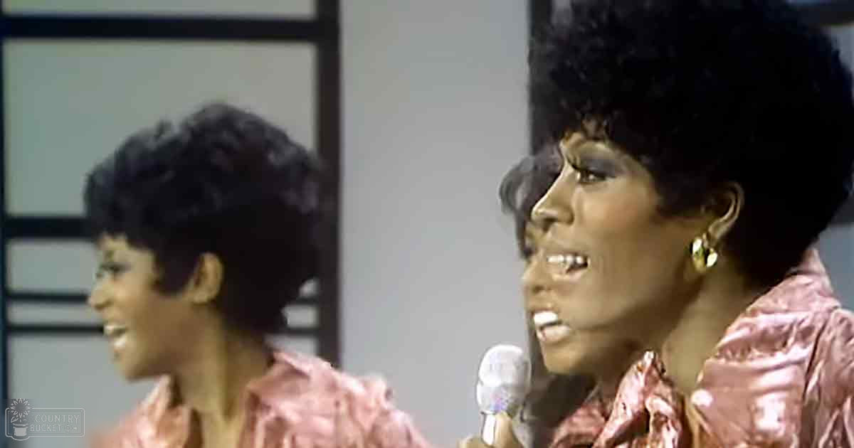 A young adult female singer wearing a shiny pink blouse stands to the left of an adult female singer also dressed in a similar pink blouse, both singing into a microphone in what appears to be a recording studio with natural light coming through a window frame.