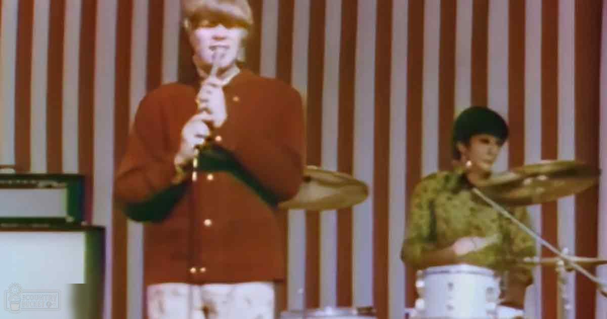 A male musician playing a flute and another man drumming indoors, with vertical red and cream striped backdrop suggesting a temporary stage setup.