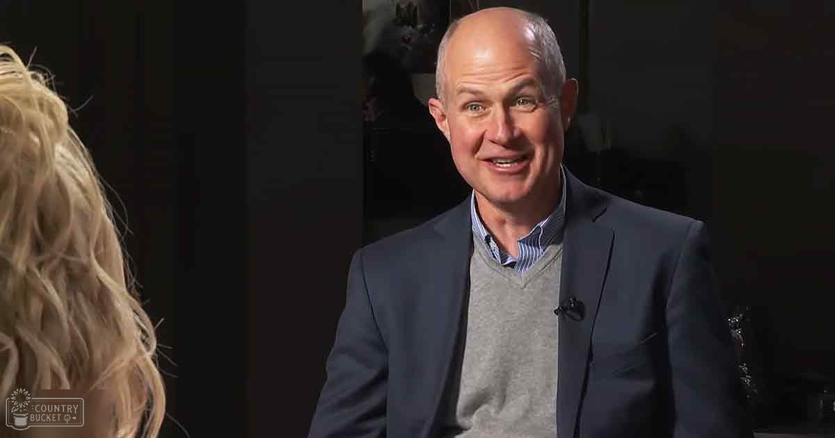 Middle-aged man in a modern, professional indoor setting wearing a dark blue blazer over a gray sweater vest with horizontal striped shirt.