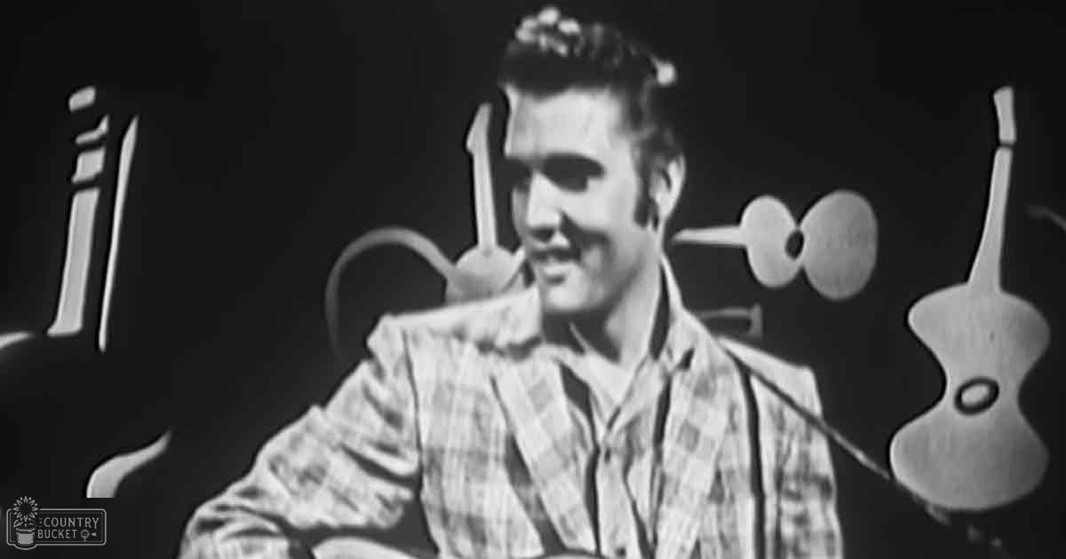 A young adult male in a plaid shirt performs on a stage, surrounded by musical instruments under focused indoor lighting.