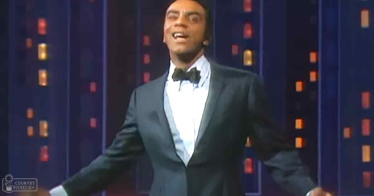 An adult male performer in a formal tuxedo with a bow tie, standing on a stage with dark blue vertical panels and small illuminated squares.
