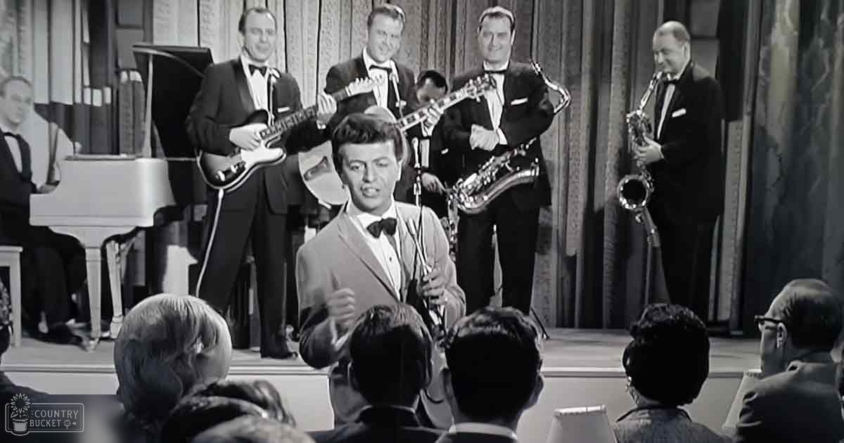 A young adult male performer in a light-colored suit jacket and white shirt with black bow tie, singing center stage with a band dressed formally in dark suits, white shirts, and black bow ties. A white grand piano is partially visible to the left.