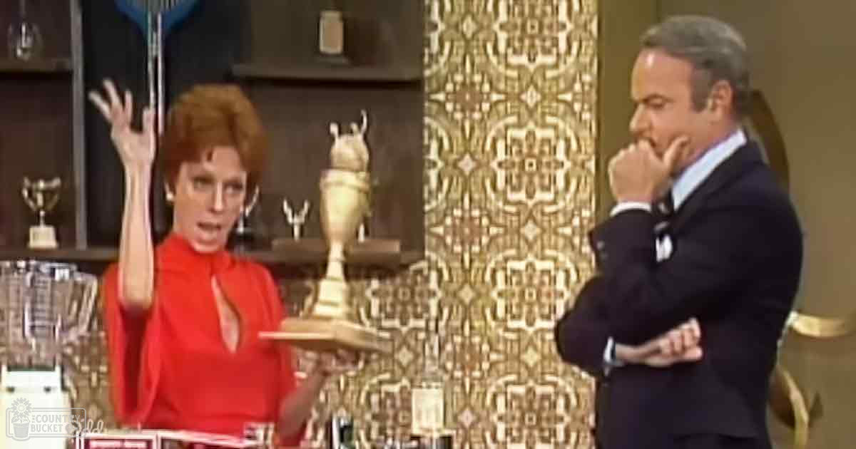 Adult female in a bright red dress with glasses, adult male in a dark suit jacket; trophy display and ornate gold background in an indoor studio setting.