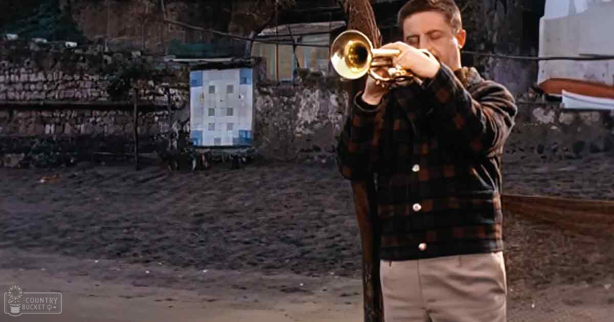 Young adult male playing a trumpet in an outdoor sandy setting with makeshift shelters in the background during daytime.